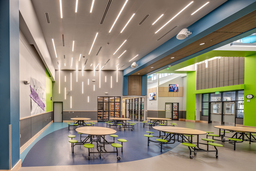 Bright modern school cafeteria with round tables, green stools, tall ceiling lights, and glass library entrance