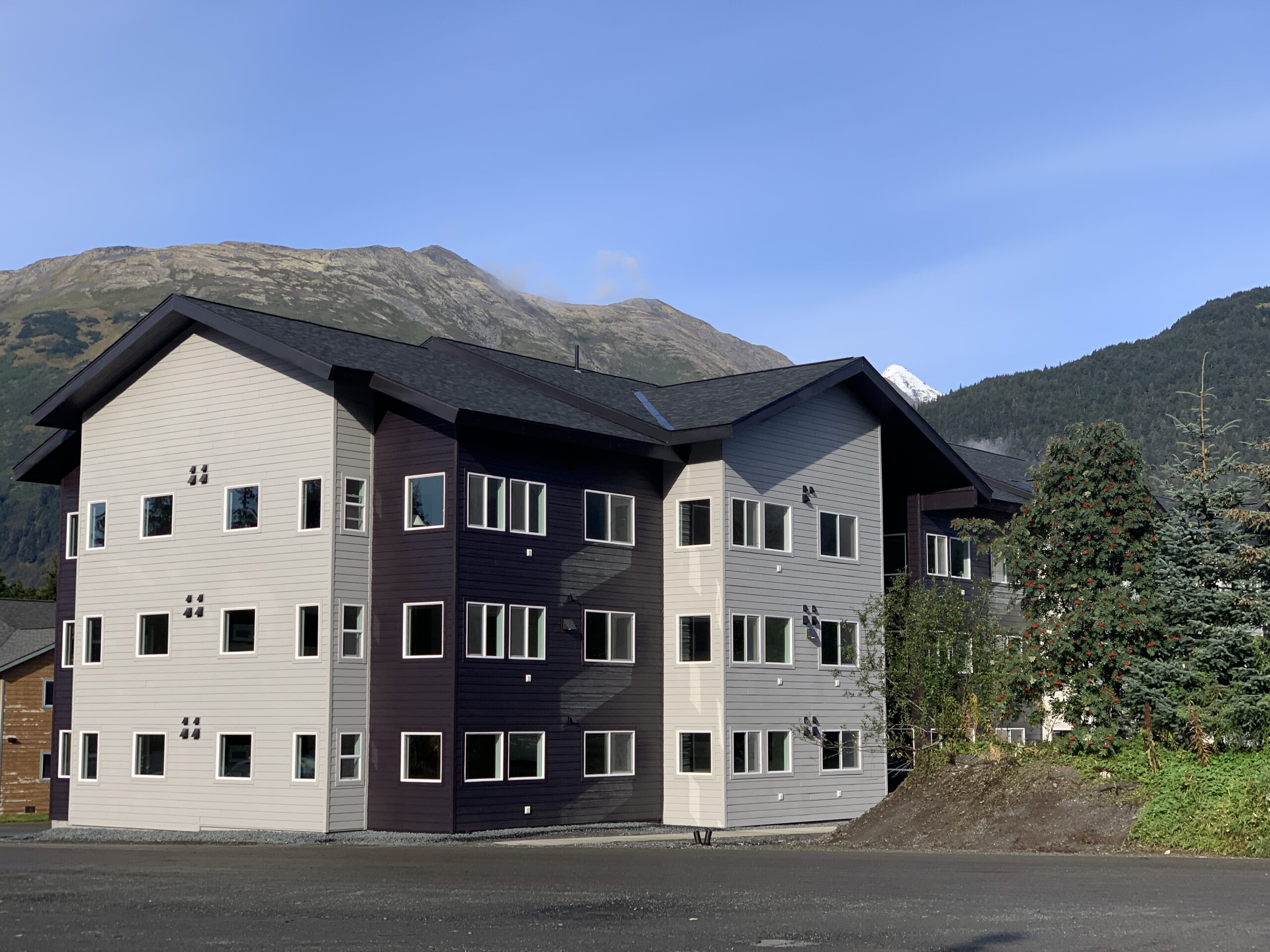 Three-story residential building with gray and dark siding, surrounded by trees and mountains in the background