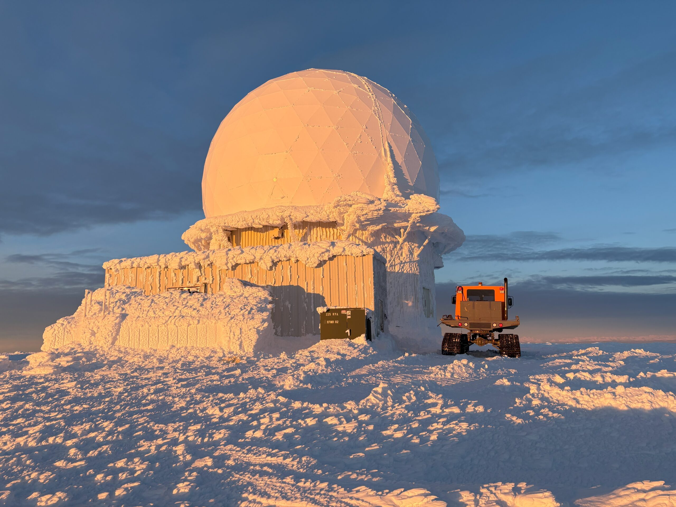 Snow-covered radar dome with an orange tracked vehicle parked in front under a golden sunset sky