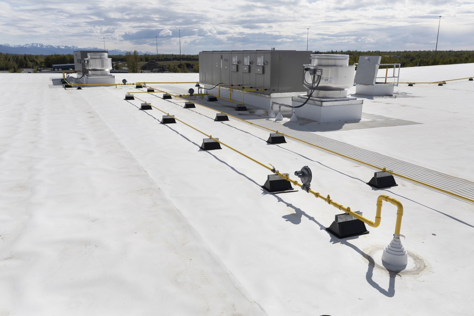 Flat commercial roof with HVAC units, ventilation fans, and yellow gas piping against a backdrop of trees and mountains