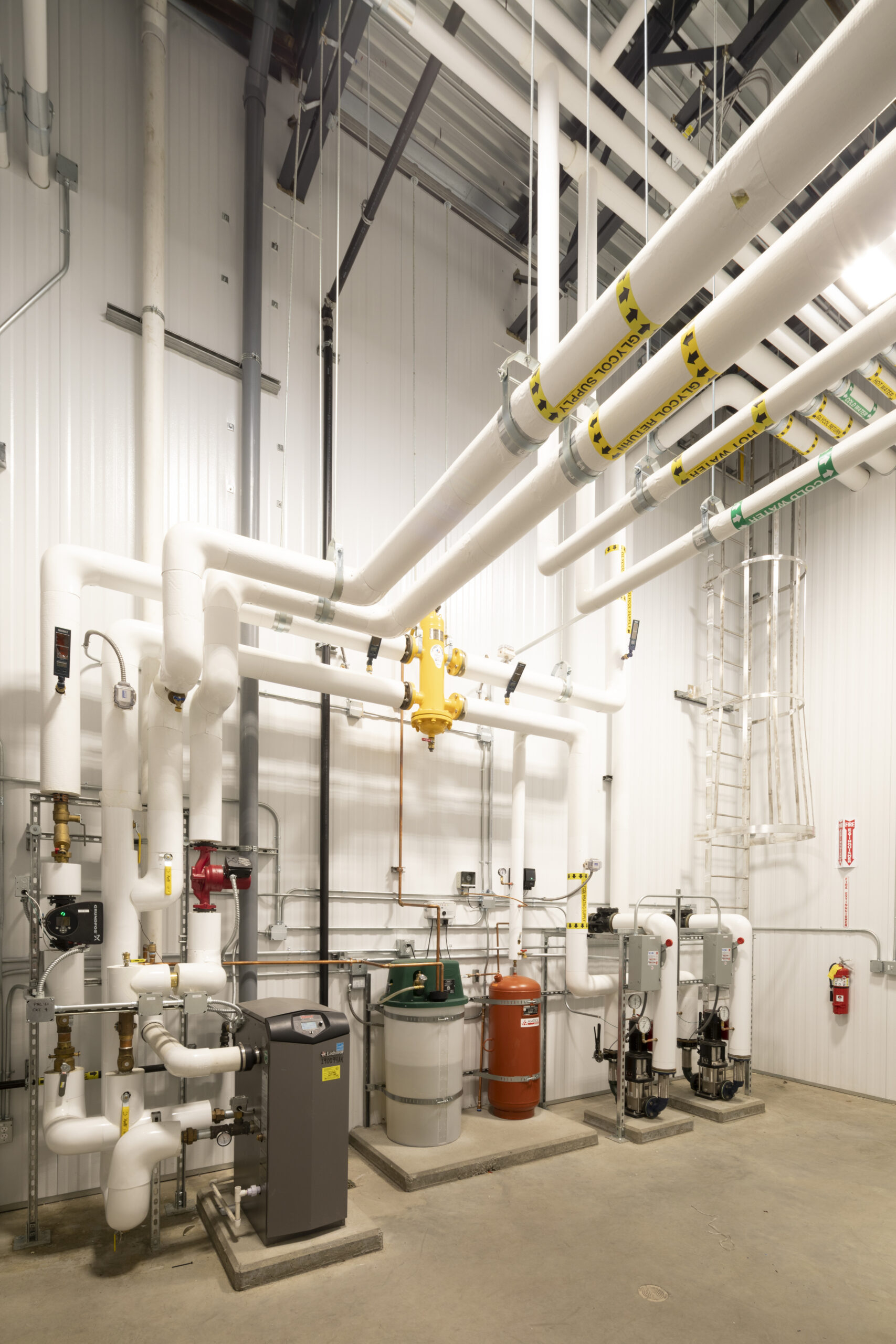 Industrial mechanical room with insulated white piping, yellow valve system, gauges, and vertical safety ladder