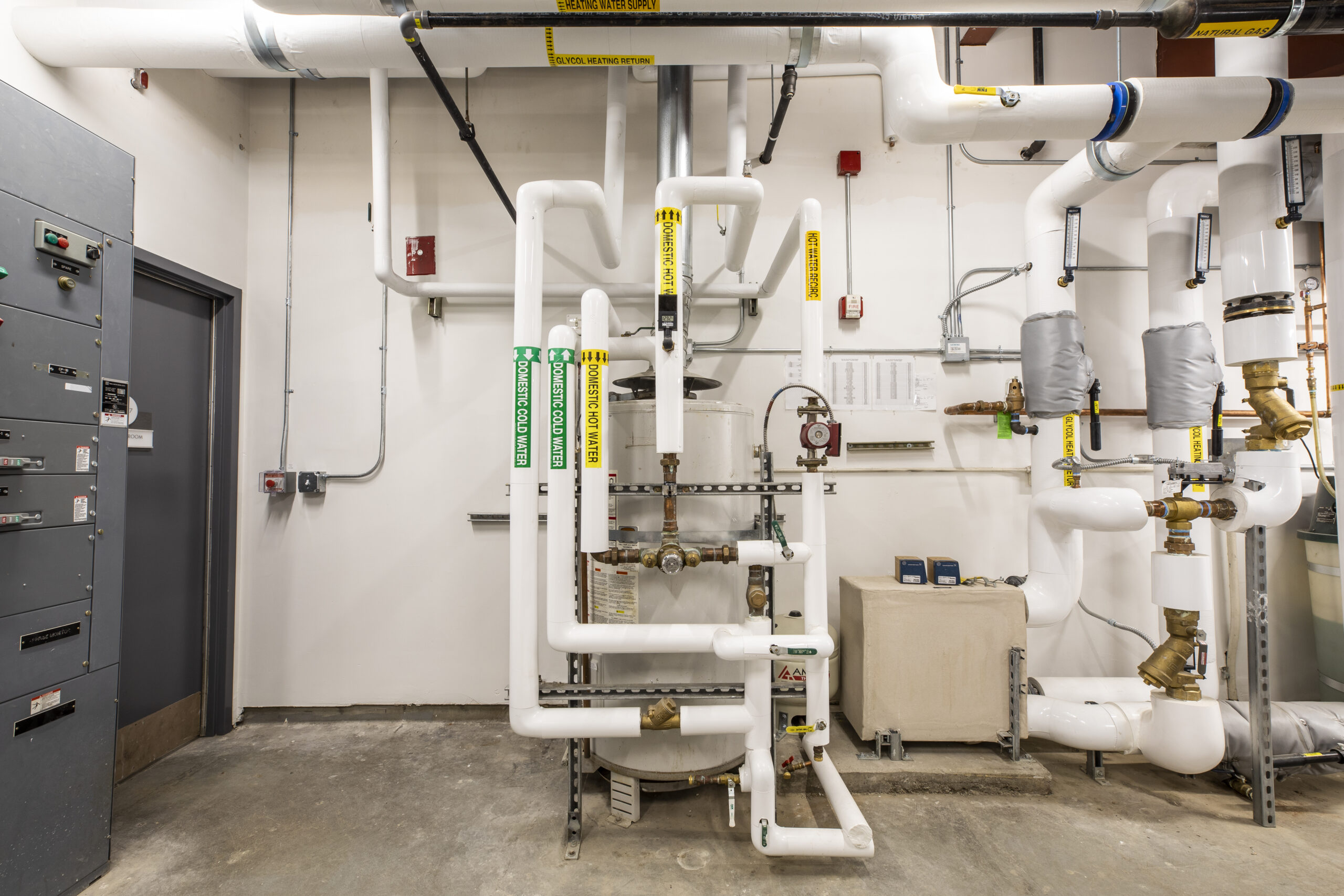 Mechanical room with insulated hot and cold water pipes, labeled valves, gauges, and electrical panels on wall