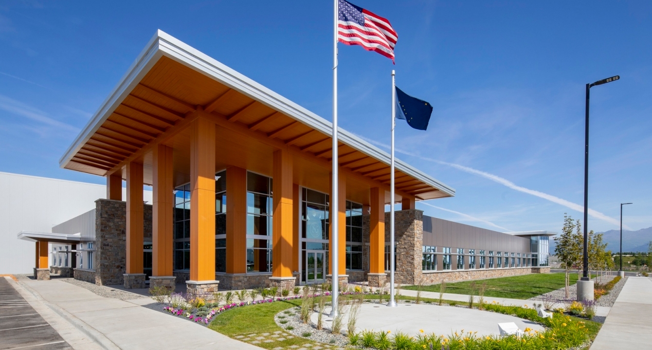 Modern building with tall glass entrance, stone columns, and U.S. and Alaska flags in front