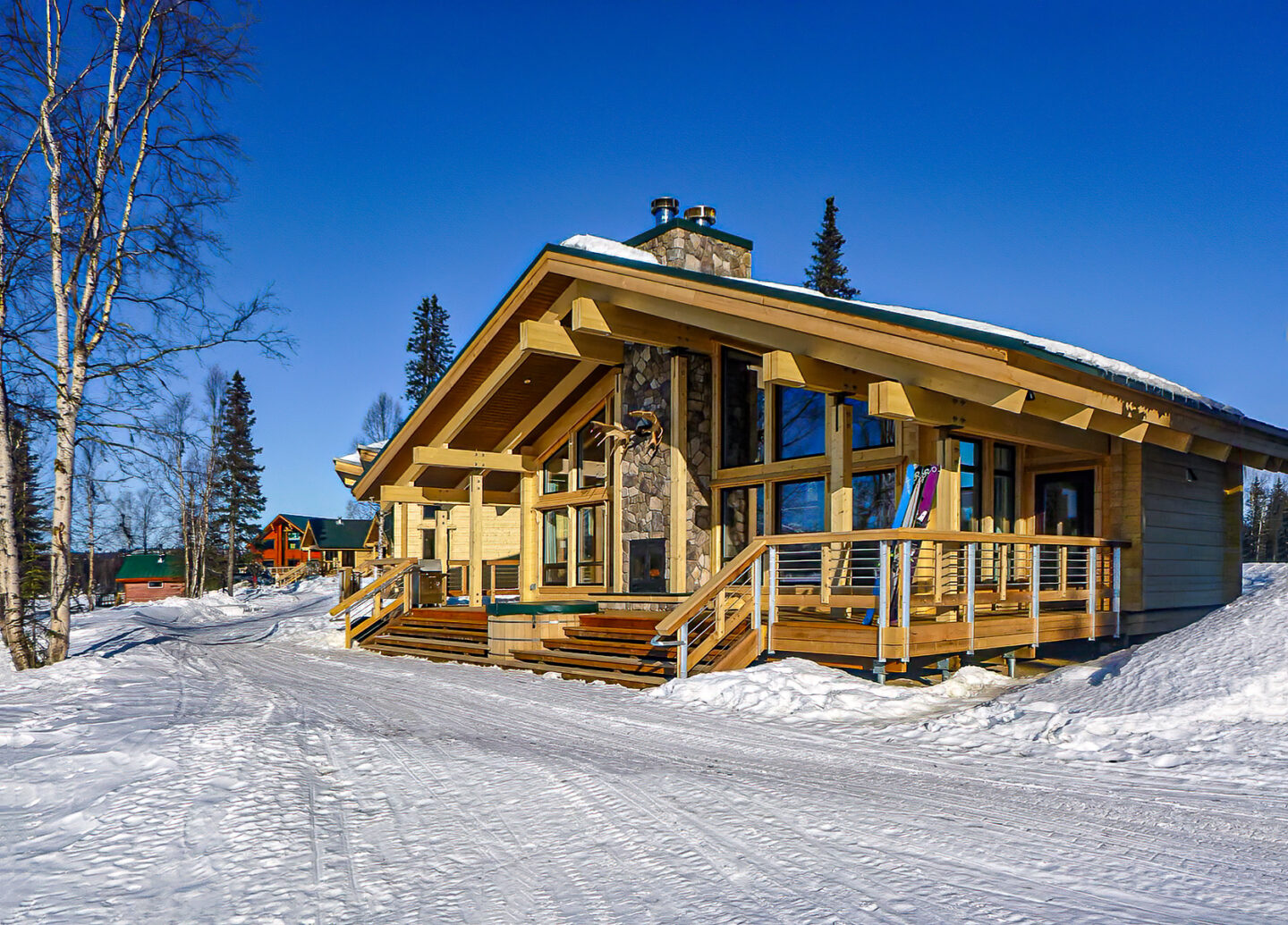 Modern wooden cabin with stone chimney and large windows, surrounded by snow and pine trees under a clear blue sky