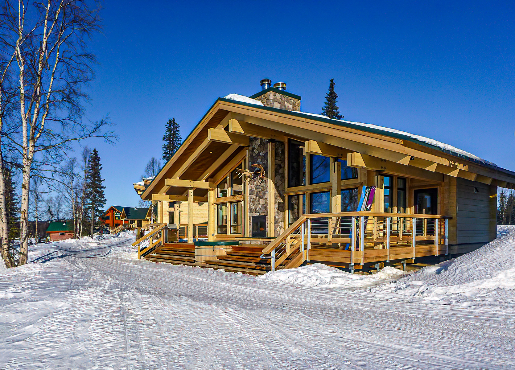 Modern wooden cabin with stone chimney and large windows, surrounded by snow and pine trees under a clear blue sky