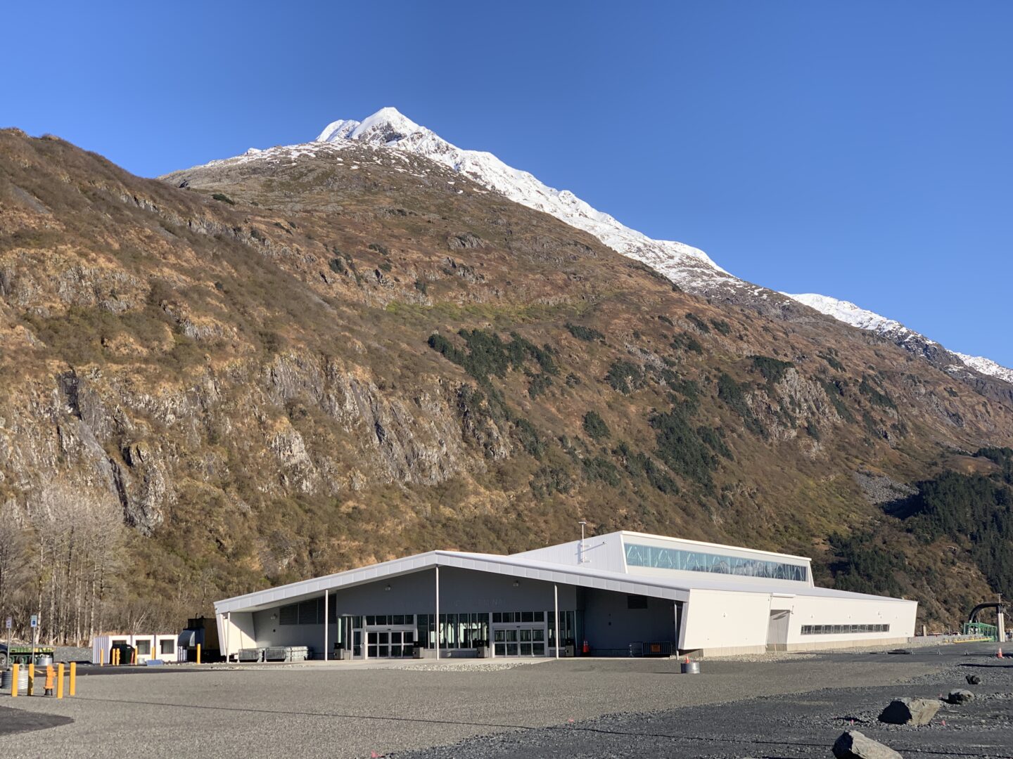Modern white building at the base of a steep mountain with snowy peak under a clear blue sky