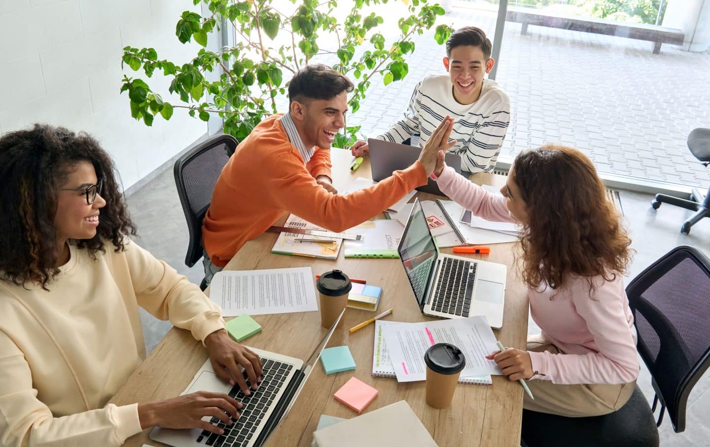 Group of students collaborating at table with laptops, giving high five in celebration